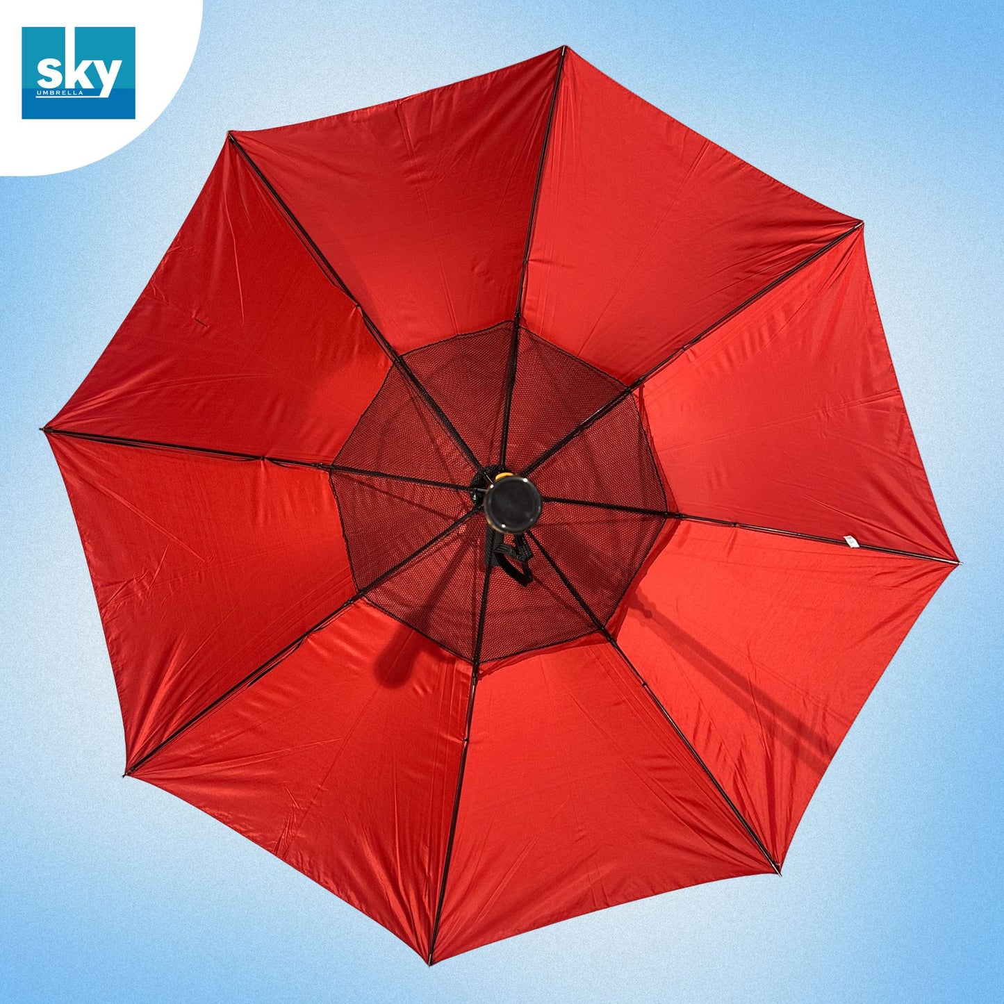 A large red Fan umbrella viewed from directly above, showing its canopy panels, ribs, and central black mesh hub against a light blue sky. 