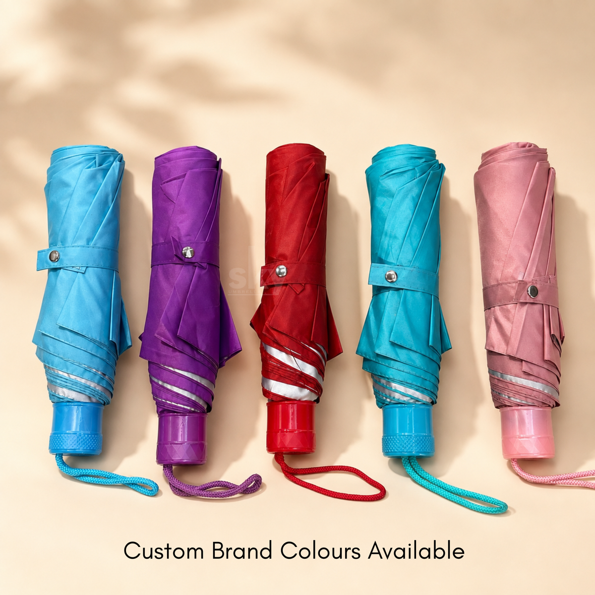 Five folded umbrellas in different colors—light blue, purple, red, teal, and pink are lined up side by side on a warm beige background with soft leaf shadows. Text at the bottom reads 'Custom Brand Colours Available'.