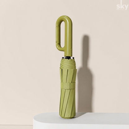 A folded green umbrella with a unique curved handle and auto open feature, standing upright on a white platform against a beige background with a soft shadow.