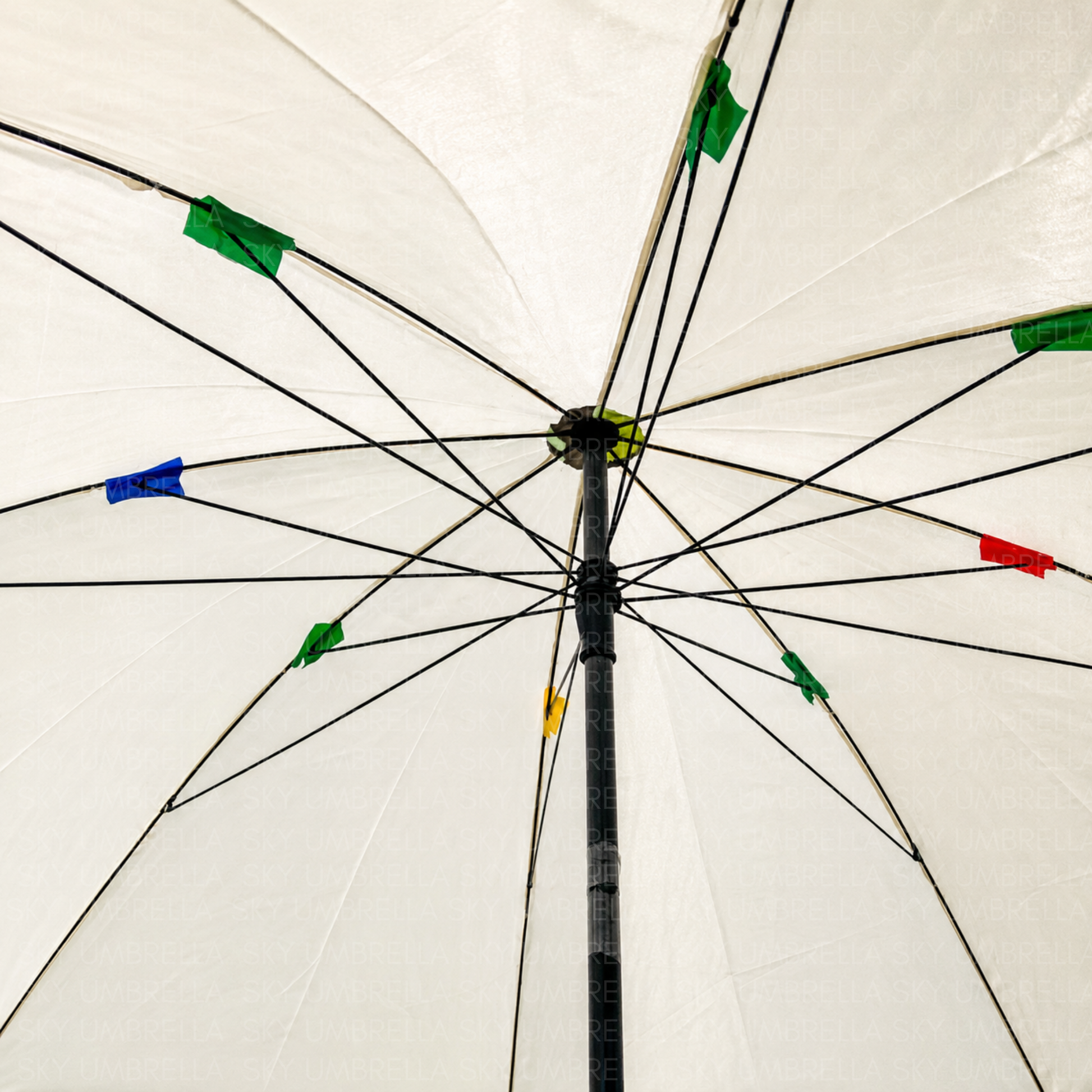 The underside of a white umbrella viewed from below, showing the black metal ribs, central hub, and small colored plastic tips in green, blue, red, yellow, and black.