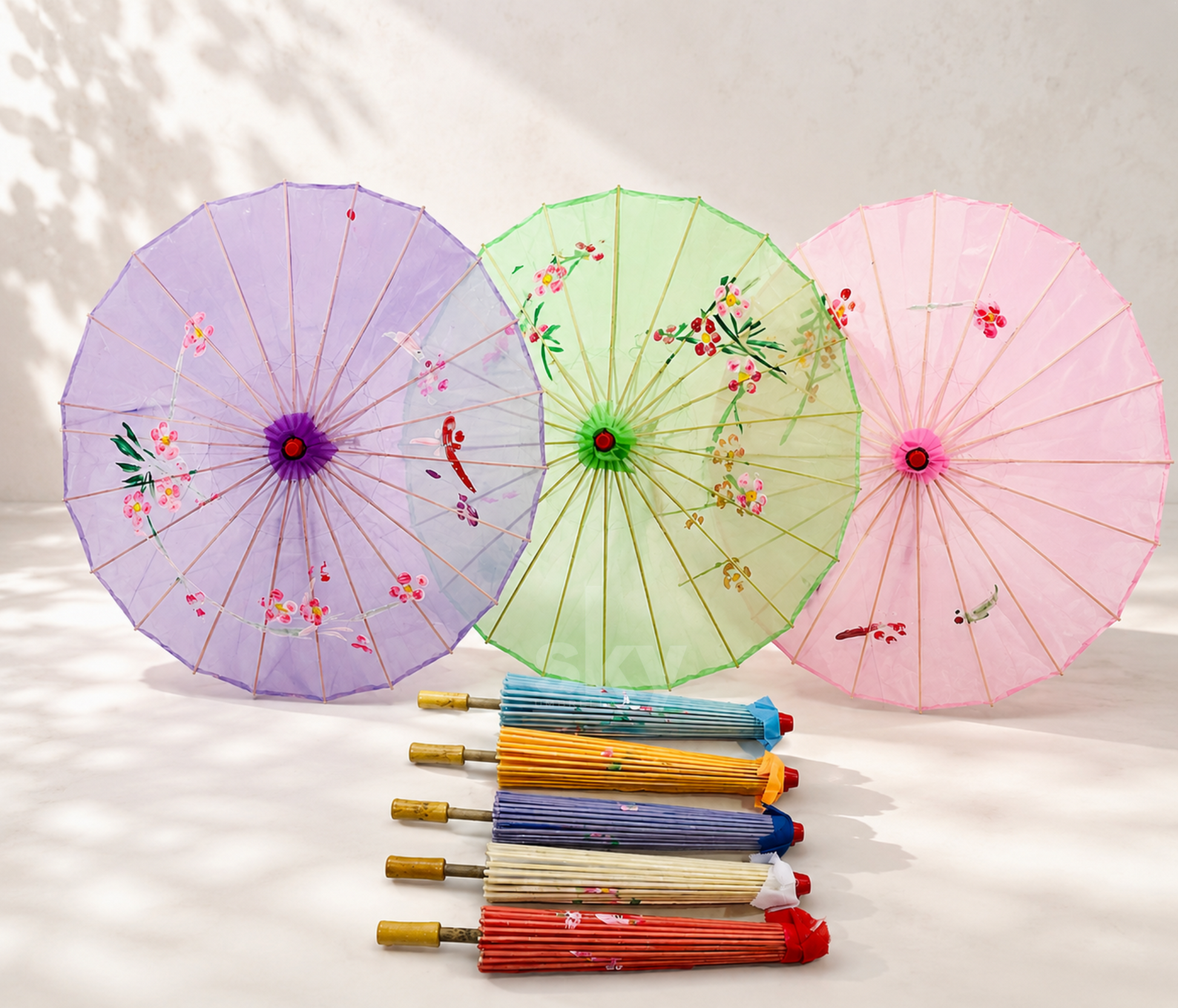 Three traditional Japanese-style paper umbrellas in purple, light green and pink, decorated with floral and bird motifs, displayed alongside a row of folded multicolored umbrellas on a light beige background.