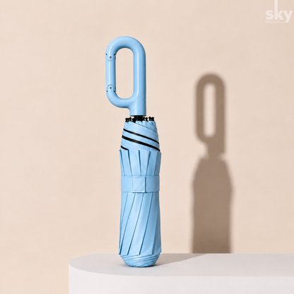 A light blue compact umbrella with a built-in carabiner handle, standing upright on a white pedestal against a beige background with a soft shadow.