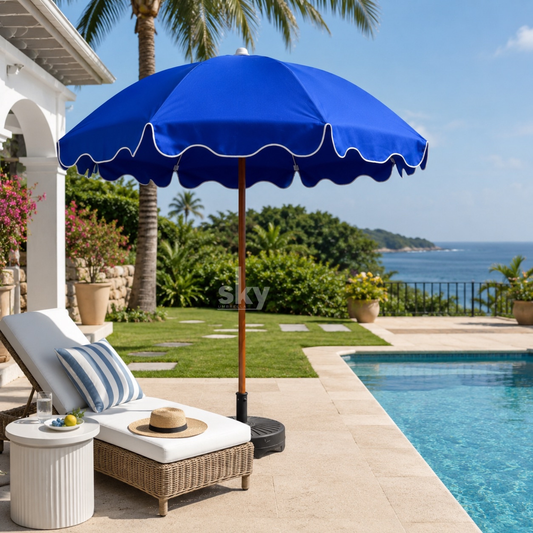 A bright blue patio umbrella with a scalloped edge stands beside a pool in a luxurious outdoor setting. A white chaise lounge with blue-striped pillows sits nearby, along with a small side table and a straw hat.