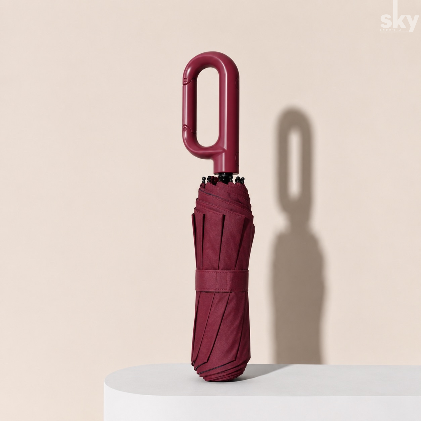 A folded maroon umbrella with a unique loop handle, standing upright on a white pedestal against a beige background with a soft shadow.