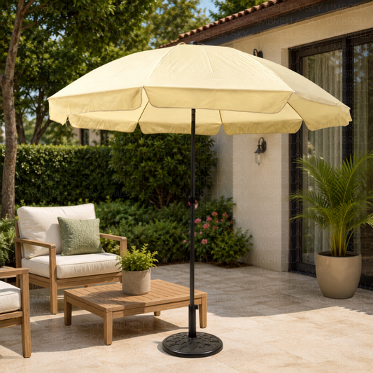A large cream patio umbrella with a black pole and base stands on a stone patio next to a wooden lounge chair, coffee table, and potted plant in front of a house.