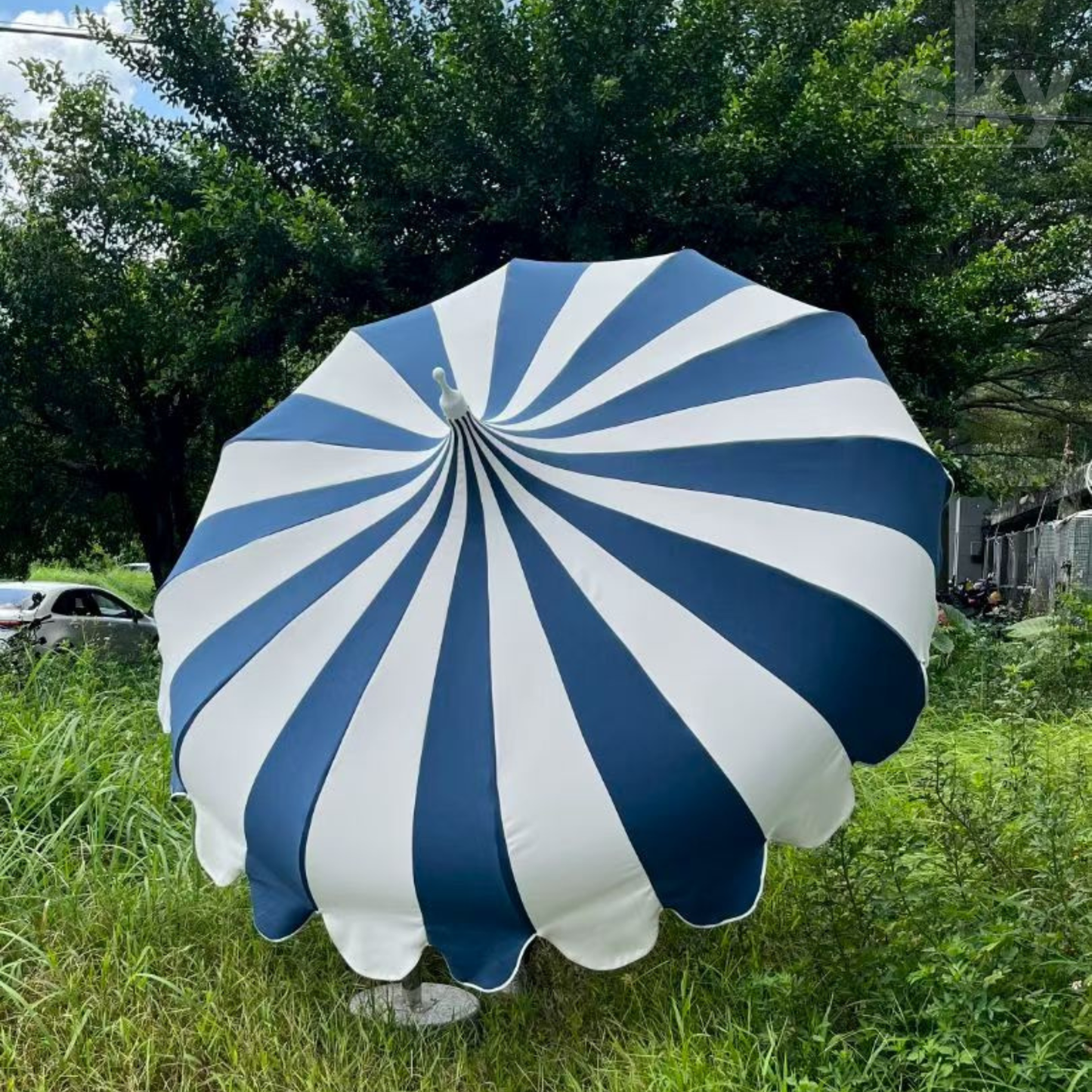A large patio umbrella with alternating navy blue and white radial stripes, set outdoors on a grassy lawn with trees in the background.