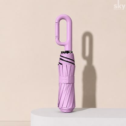 A compact, folded pink umbrella with a carabiner-style handle stands upright on a white cylindrical pedestal against a warm beige background.