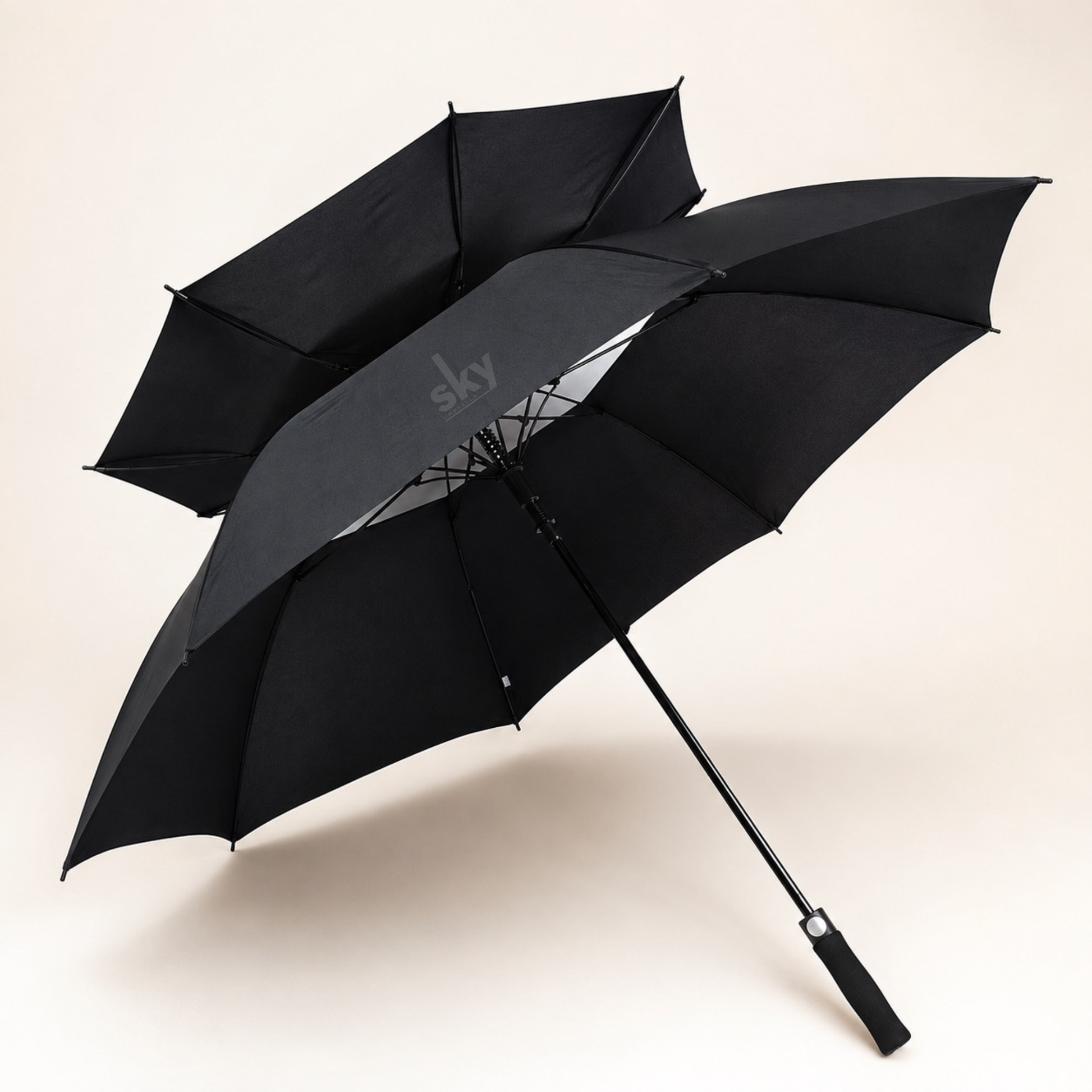 A black double umbrella with two overlapping canopies and a long handle, photographed on a clean light beige studio background. The inner canopy shows a subtle grey promotional 'sky Umbrella' logo.