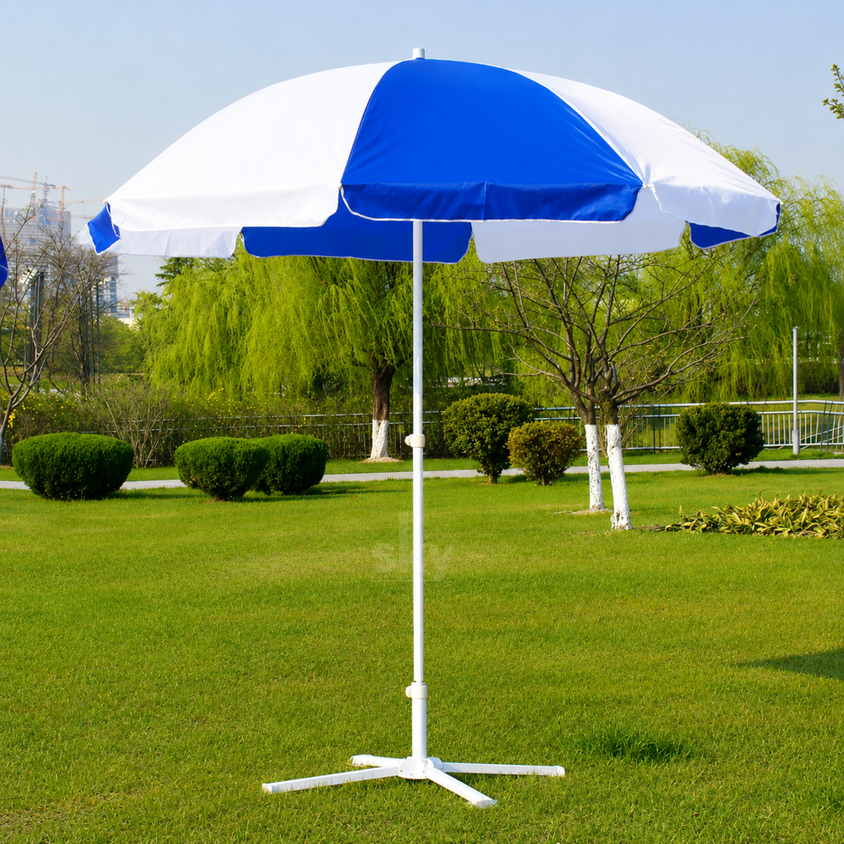A blue and white patio umbrella with a white tripod base, set up on a green grass lawn in a park with trees in the background.