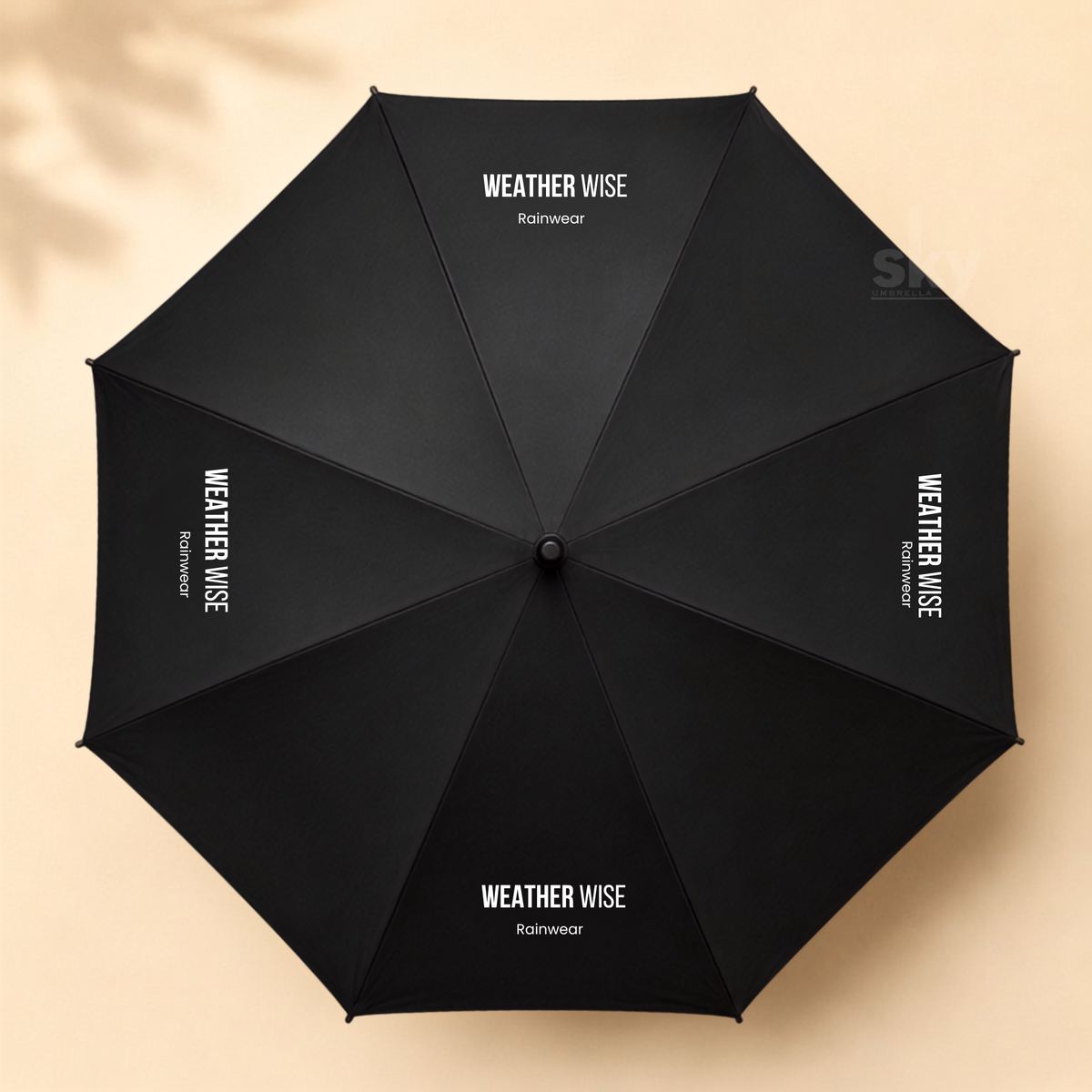 A black open umbrella viewed from above, with white 'WEATHER WISE Rainwear' branding printed on multiple panels, set against a warm beige background with soft leaf shadows.