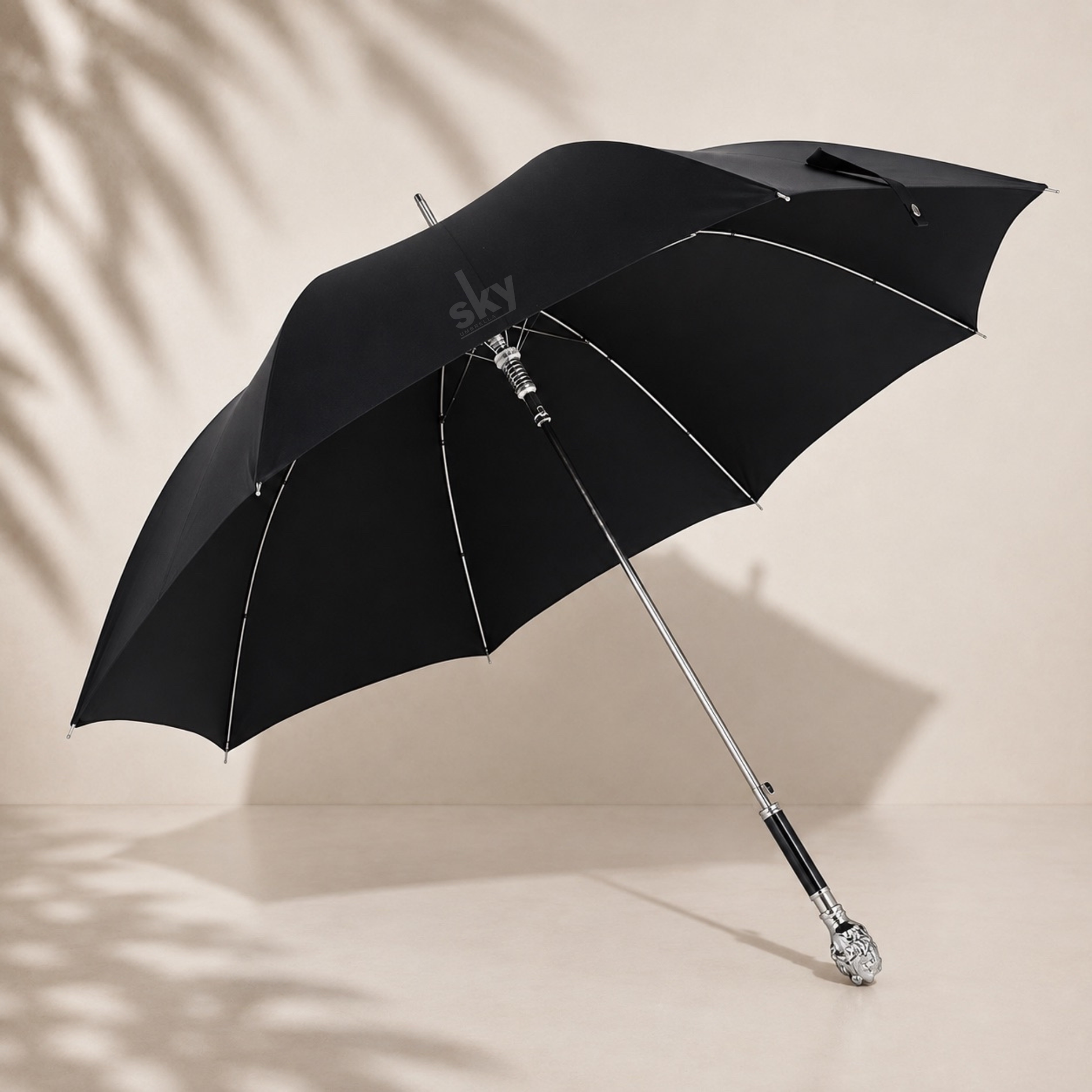 A black open umbrella with silver metal ribs and unique lion handle, photographed on a warm beige studio background with soft palm leaf shadows.